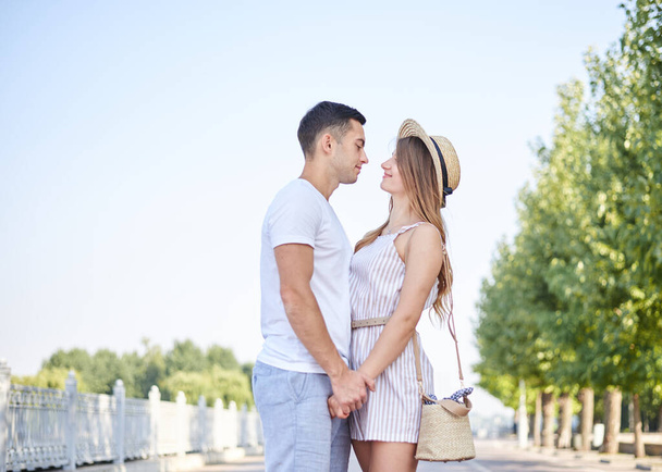 Handsome brunette man, wearing white t-shirt, blue shorts, is hugging pretty blond woman in light straw hat and stripy summer overall. Young couple in love, embracing, looking at each other, smiling. - 写真・画像