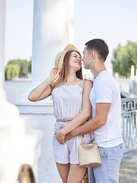 Handsome man, wearing white t-shirt, blue shorts, hugging from behind pretty woman in light straw hat and stripy summer overall. Young couple in love, embracing, looking at each other, kissing. - Foto, afbeelding