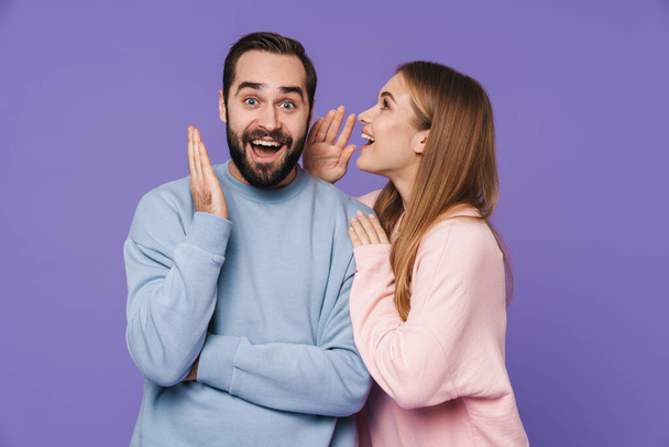 Image of emotional surprised young loving couple isolated over purple background gossiping talking with each other. - Fotoğraf, Görsel