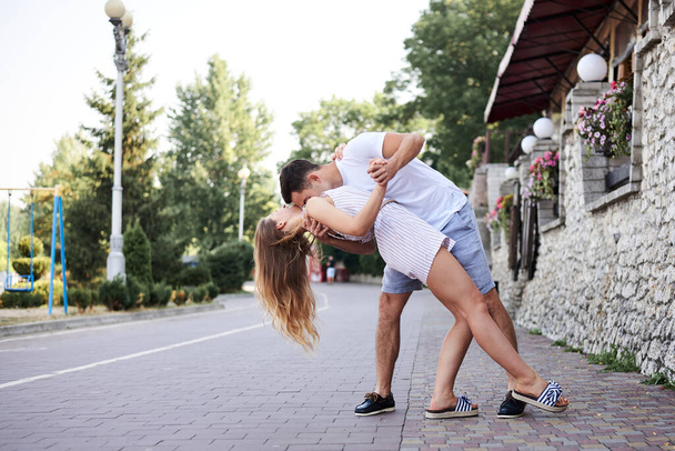 Young couple in love hugging kissing near stone wall in resort town. Pretty blond woman, wearing stripy short overall and brunette man in white t-shirt and blue shorts on romantic date. Relationship - Photo, Image