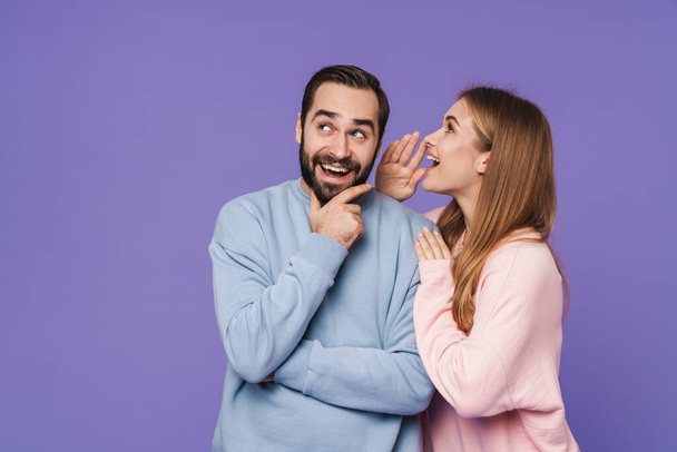 Picture of emotional positive young loving couple isolated over purple background gossiping talking with each other. - Photo, Image