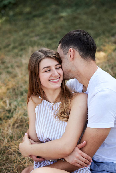 Young couple in love sitting on green yellow grass lawn hugging embracing kissing. Blond woman wearing stripy short overall and brunette man in white t-shirt blue shorts on romantic date. Relationship - Foto, immagini
