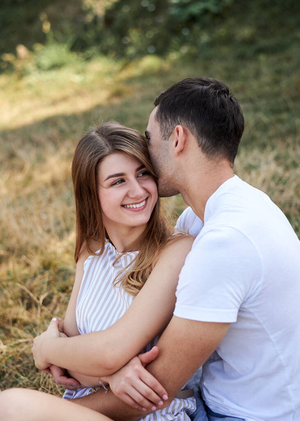 Young couple in love sitting on green yellow grass lawn hugging embracing kissing. Blond woman wearing stripy short overall and brunette man in white t-shirt blue shorts on romantic date. Relationship - Foto, Imagem