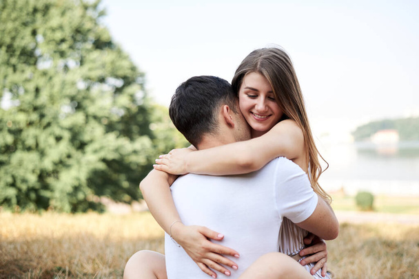 Young couple in love sitting on green yellow grass lawn hugging embracing kissing. Blond woman wearing stripy short overall and brunette man in white t-shirt blue shorts on romantic date. Relationship - Фото, изображение