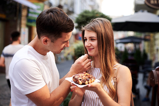 Romantic couple on date eating tasty belgian waffle with chocolate and caramel. Young blond woman, feeding brunette man with yummy desert in summer in the morning. Nutritious breakfast, snack. - Photo, Image