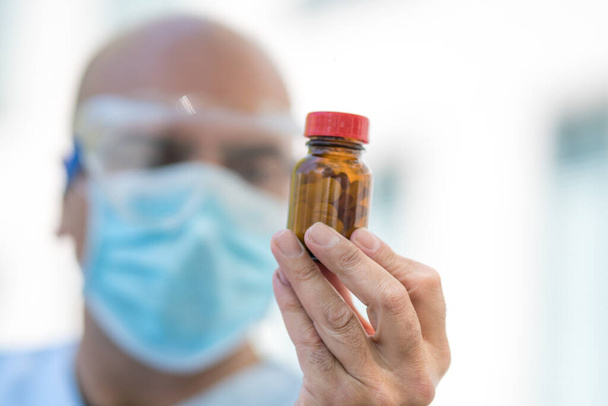 Πορτρέτο ενός εκτός εστίασης γιατρού φορώντας λευκό παλτό, χειρουργική μάσκα και γυαλιά ιατρικής ασφάλειας κρατώντας ένα μπουκάλι χάπια στο χέρι του. Επιλεκτική εστίαση - Φωτογραφία, εικόνα