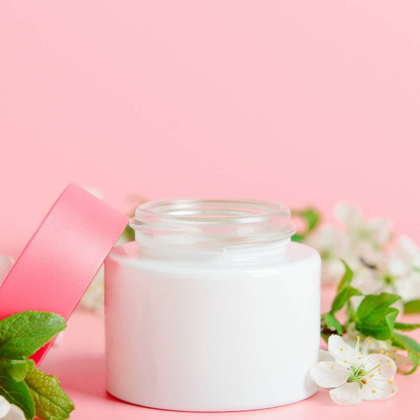 Face cream in white jar and pink cover on a pink background with white flowers. Concept natural cosmetics, organic beauty - Photo, image
