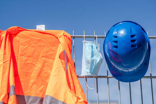 Arbeitssicherheit und Schutz vor widrigen Arbeitsbedingungen. Blauer Bollenhut, reflektierende Weste und chirurgische Schutzmaske hängen auf einem Gerüst - Foto, Bild