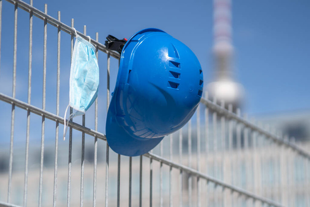 Occupational safety and protection against adverse conditions at work. Blue hard hat and protective surgical mask hanging on scaffolding. On background out of focused Berlin television tower - Photo, Image