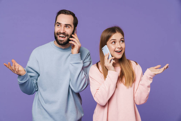 Foto van gelukkig optimistisch liefdevol paar geïsoleerd over paarse achtergrond praten door mobiele telefoons. - Foto, afbeelding