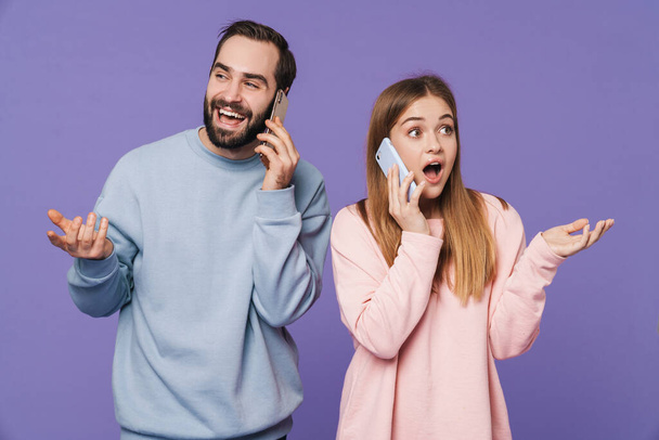 Picture of happy optimistic loving couple isolated over purple background talking by mobile phones. - Zdjęcie, obraz