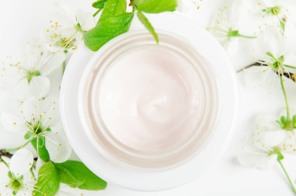 Face cream in white jar on a white background with small flowers of an apple tree. Concept natural cosmetics, organic beauty, flower arrangement. Top view, flat lay - Photo, image