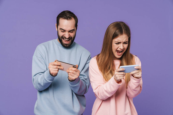 Image of excited nervous loving couple isolated over purple background play games by mobile phones. - Zdjęcie, obraz