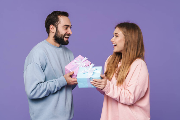 Image of a happy young loving couple isolated over purple wall background give a gifts for each other. - Zdjęcie, obraz