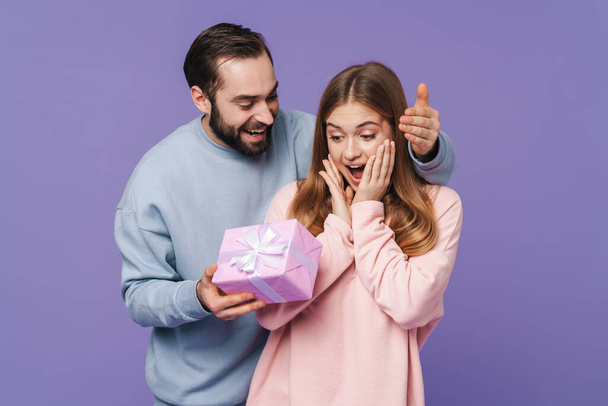 Image of a happy young man give a gift for his girlfriend isolated over purple wall background. - Photo, Image