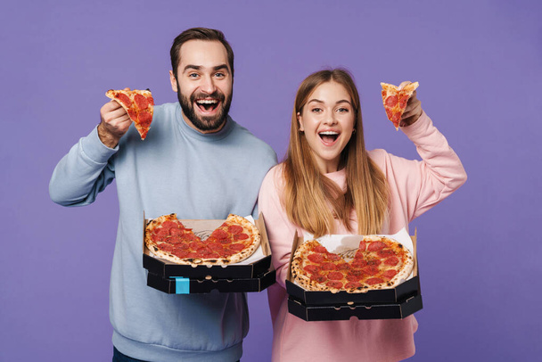 Photo of a positive emotional loving couple friends isolated over purple wall background eat pizza. - 写真・画像