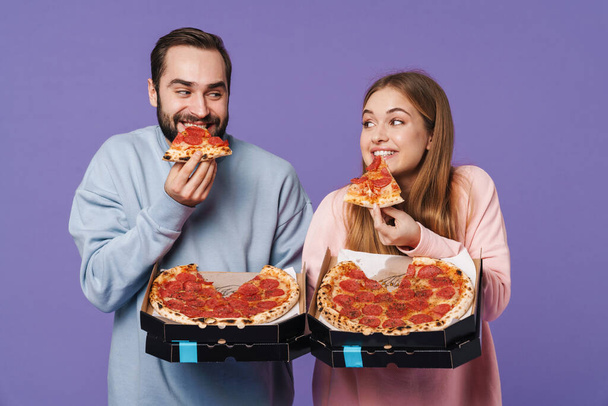 Fotografie pozitivního emocionální milující pár přátel izolovaných přes fialové stěny pozadí jíst pizzu. - Fotografie, Obrázek