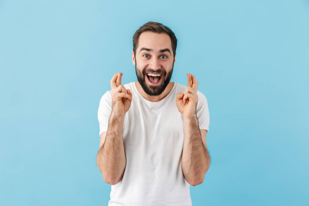 Portrait of a young cheerful excited bearded man wearing t-shirt standing isolated over blue background, holding fingers crossed for good luck - Zdjęcie, obraz