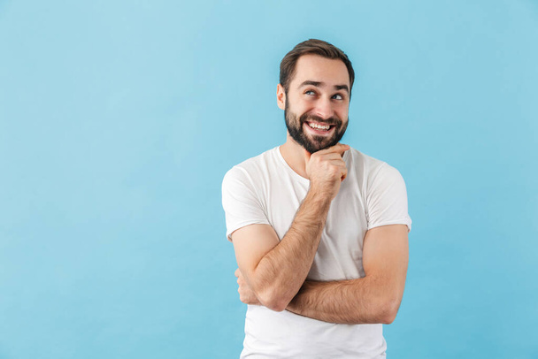 Egy fiatal, mosolygós, elmélkedő, szakállas férfi portréja, aki pólóban áll elszigetelve a kék háttér felett. - Fotó, kép