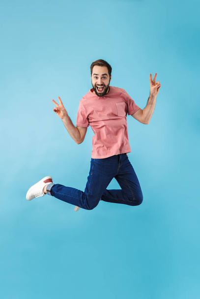 Imagem do jovem bonito animado feliz homem pulando isolado sobre fundo parede azul fazer gesto de paz
. - Foto, Imagem