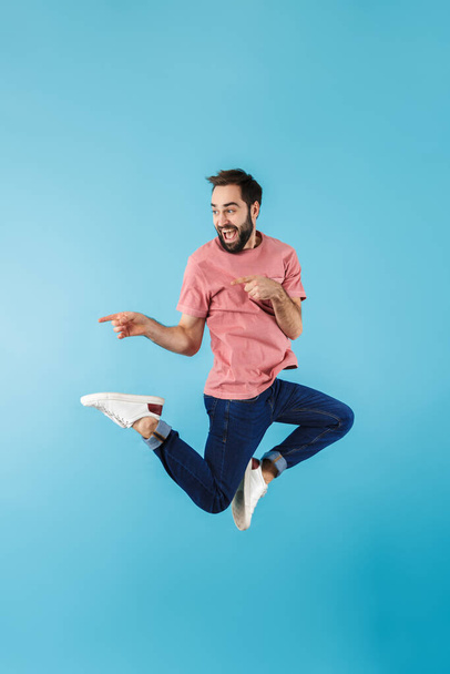 Full length of a young cheerful excited bearded man wearing t-shirt jumping isolated over blue background, pointing away at copy space - Fotó, kép