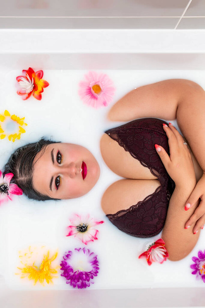 woman wearing sensual red lingerie on a milky bathtub with flowers. - Photo, image