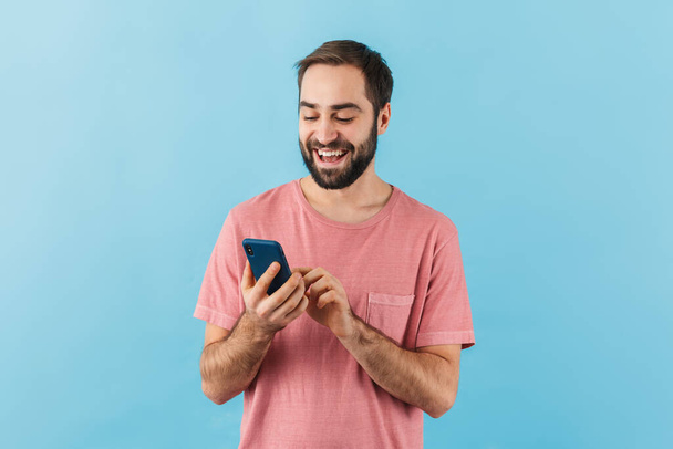 Portrait of a young cheerful excited bearded man wearing t-shirt standing isolated over blue background, using mobile phone - Foto, immagini