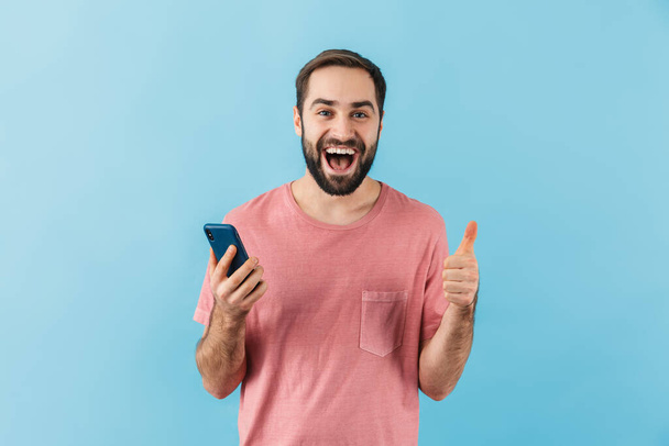 Portrait of a young cheerful excited bearded man wearing t-shirt standing isolated over blue background, using mobile phone, showing thumbs up - Photo, Image