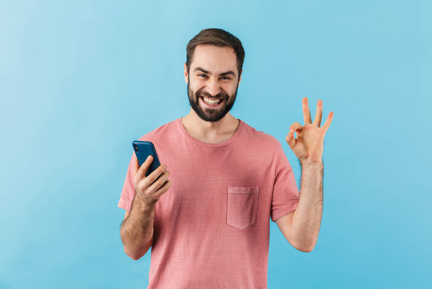 Portrait of a young cheerful excited bearded man wearing t-shirt standing isolated over blue background, using mobile phone, showing ok - 写真・画像