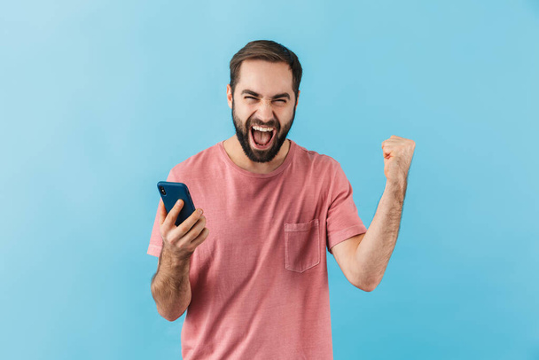 Portrait of a young cheerful excited bearded man wearing t-shirt standing isolated over blue background, using mobile phone, celebrating success - Foto, imagen