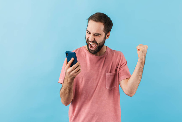 Portrait of a young cheerful excited bearded man wearing t-shirt standing isolated over blue background, using mobile phone, celebrating success - Фото, изображение