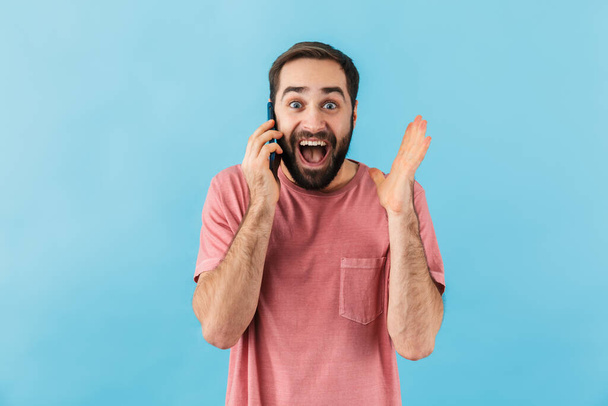 Image of young handsome surprised positive man isolated over blue wall background talking by mobile phone. - Foto, afbeelding