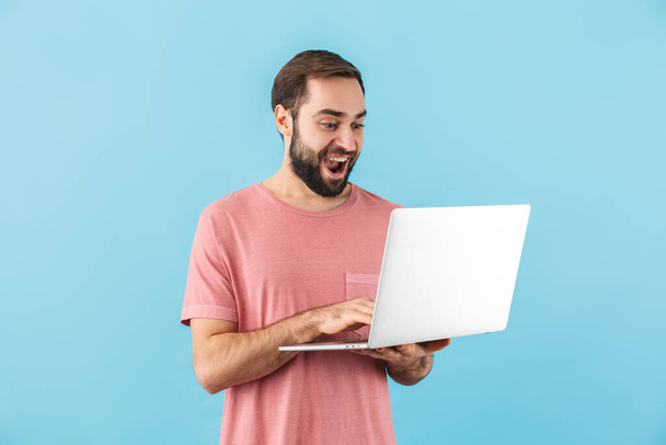 Portrait of a young cheerful excited bearded man wearing t-shirt standing isolated over blue background, holding laptop computer - Photo, Image