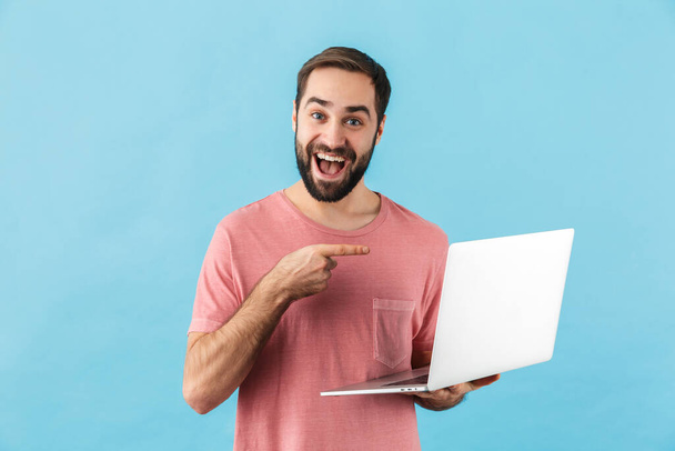 Portrait of a young cheerful excited bearded man wearing t-shirt standing isolated over blue background, pointing finger at laptop computer - Foto, Imagem