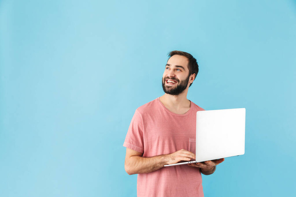 Portrait of a young cheerful excited bearded man wearing t-shirt standing isolated over blue background, holding laptop computer, looking up - Photo, Image