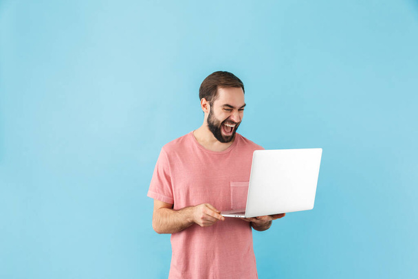 Portrait of a young cheerful excited bearded man wearing t-shirt standing isolated over blue background, holding laptop computer - Foto, immagini