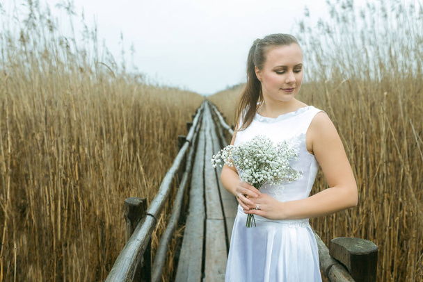Ritratto di sposa in abito da sposa lungo bianco con mazzolino bianco sullo sfondo delle canne, lago e passerella in legno
  - Foto, immagini