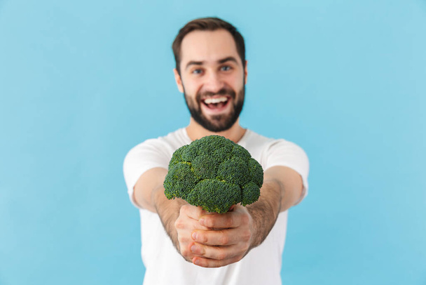 Portrait of a young cheerful excited bearded man wearing t-shirt standing isolated over blue background, showing broccoli - Φωτογραφία, εικόνα