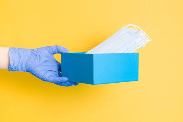eine Hand in einem blauen Einweghandschuh hält eine blaue Schachtel mit medizinischen Schutzmasken, gelbem Hintergrund, Kopierschutz - Foto, Bild