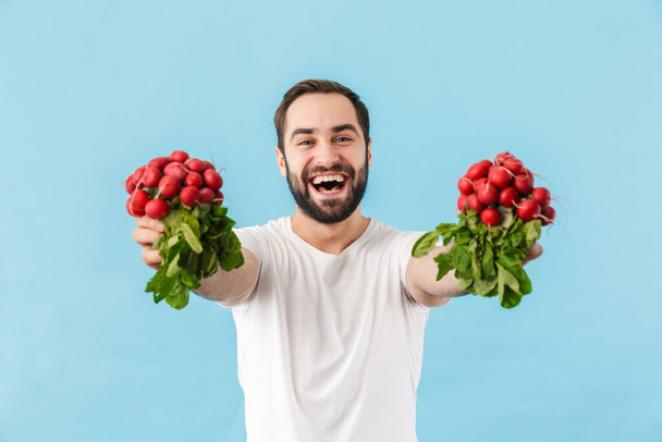 Portrait of a young cheerful excited bearded man wearing t-shirt standing isolated over blue background, showing fresh redish - 写真・画像