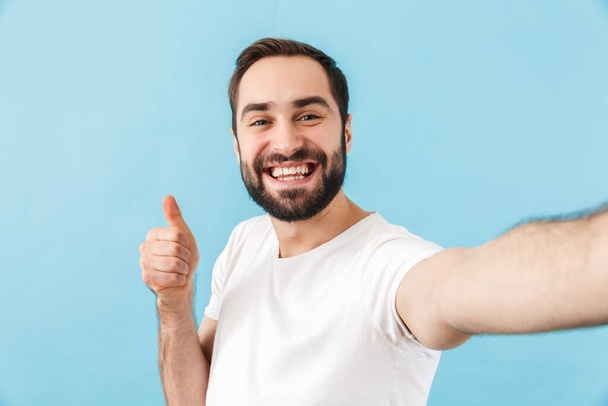 Image of young happy cheerful man isolated over blue wall background take a selfie by camera showing thumbs up. - Valokuva, kuva