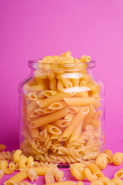 One glass jar with variety of uncooked golden wheat pasta on minimal pink background, angle view macro - Valokuva, kuva