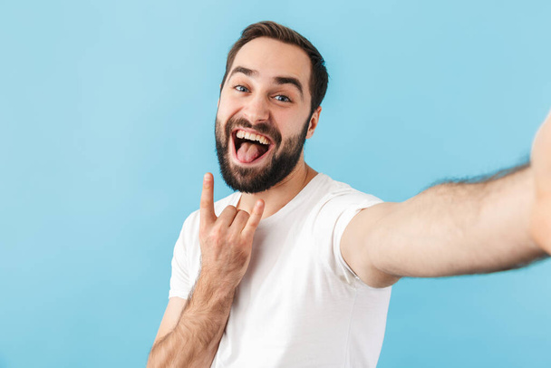 Image of young happy man isolated over blue wall background take a selfie by camera showing rock gesture. - Valokuva, kuva