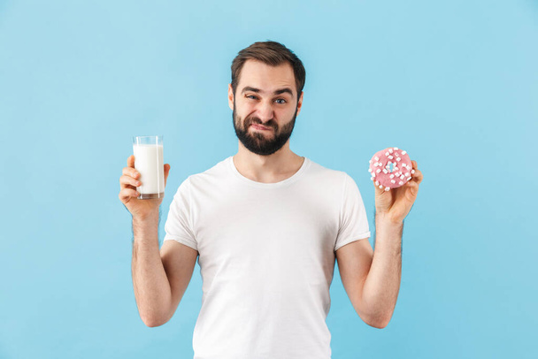 Portrait of a young angry bearded man wearing t-shirt standing isolated over blue background, showing glass of milk and a donut - Foto, immagini