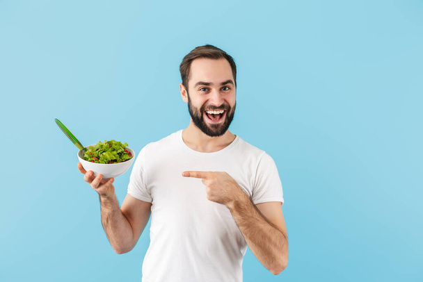 Handsome cheerful young bearded man wearing t-shirt standing isolated over blue background, holding bowl with fresh salad, pointing finger - Fotografie, Obrázek