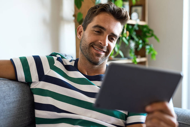 Smiling young man sitting on his living room sofa at home and using a digital tablet - Foto, Bild
