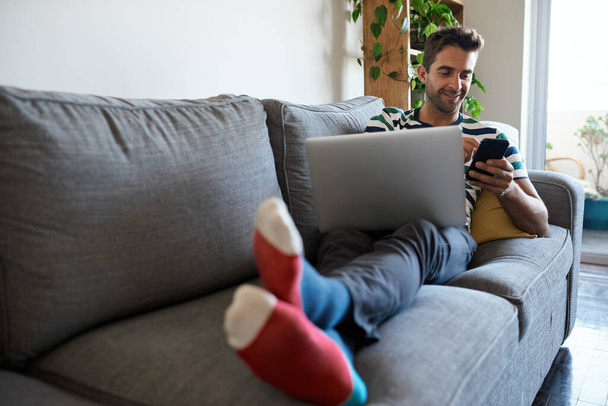 Smiling young man relaxing on his living room sofa sending a text on his cellphone and using a laptop - Φωτογραφία, εικόνα
