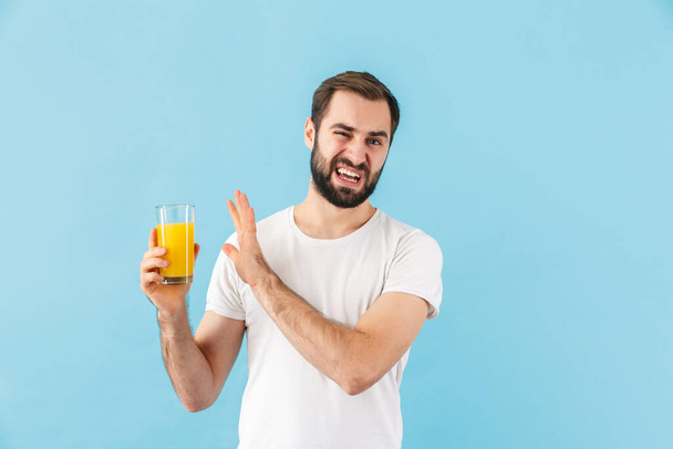 Handsome disappointed young bearded man wearing t-shirt standing isolated over blue background, showing glass of fresh orange juice - Фото, изображение