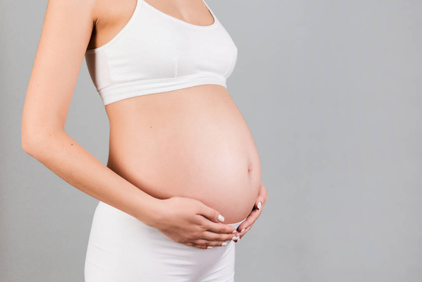 Beyaz iç çamaşırlı hamile bir kadının gri arka planda karnına dokunduğu görüntüler. Müstakbel anne bebeğini bekliyor. Boşluğu kopyala. - Fotoğraf, Görsel