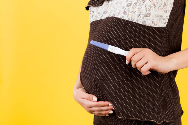 Blisko kobiety w ciąży w brązowej piżamie z dodatnim wynikiem testu ciążowego na żółtym tle. Koncepcja macierzyństwa. Przestrzeń kopiowania. - Zdjęcie, obraz
